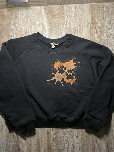 Vollständige Vorderansicht eines schwarzen Sweatshirts mit minimalistischen Bleach Art Spritzern und zwei Hundepfotenabdrücken auf Herzhöhe