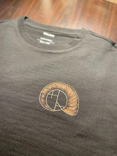 Detailaufnahme des handgezeichneten Künstlerlogos in Muschelform auf der Vorderseite des T-Shirts