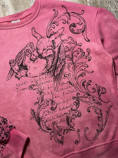 Schräge Vorderansicht des rosa handbemalten Sweatshirts mit geflügeltem Hasen und schwarzen Farbtropfen am Ärmel