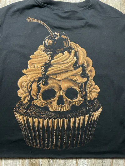 Nahaufnahme eines handgestalteten Bleach-Art-Cupcakes mit Totenkopf, Sahne und Kirsche auf einem schwarzen T-Shirt