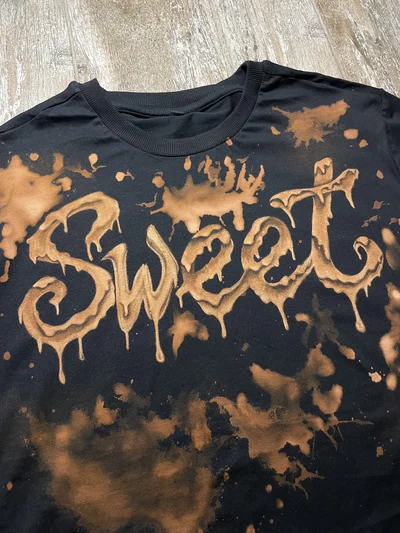 Nahaufnahme des handgestalteten SWEET-Schriftzugs und der Bleichtropfen auf der Vorderseite eines schwarzen T-Shirts