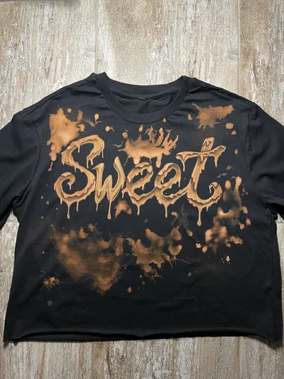 Vollständige Vorderansicht eines schwarzen Cropped-T-Shirts mit handgestaltetem SWEET-Schriftzug und Bleichtropfen