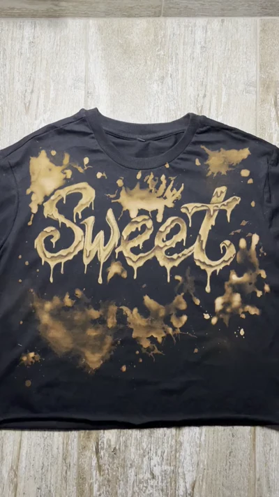 Video der Vorderseite eines schwarzen Cropped-T-Shirts mit handgestaltetem SWEET-Schriftzug und fließenden Bleichtropfen